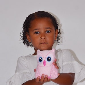 May include: A young girl with curly brown hair is holding a pink plush owl toy with embroidered blue eyes. The owl has a pink beak and pink stitching around the edges.