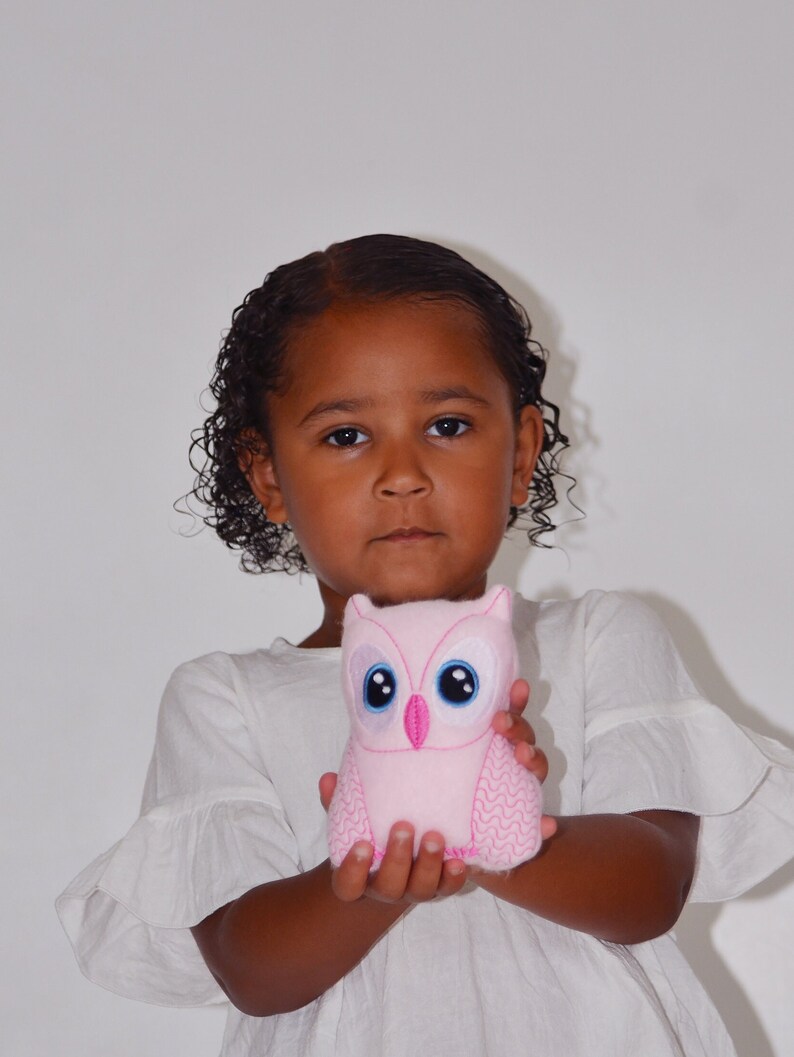 May include: A young girl with curly brown hair is holding a pink plush owl toy with embroidered blue eyes. The owl has a pink beak and pink stitching around the edges.