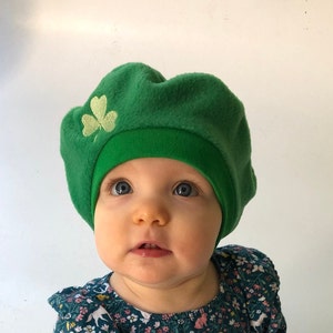 May include: A young child wearing a green fleece beret with a shamrock embroidered on the front. The beret has a green band around the bottom. The child is wearing a floral print dress.