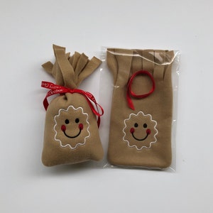 May include: Two brown felt gingerbread man ornaments with smiling faces. One ornament is in a bag with a red ribbon tied around it. The other ornament is in a clear plastic bag.