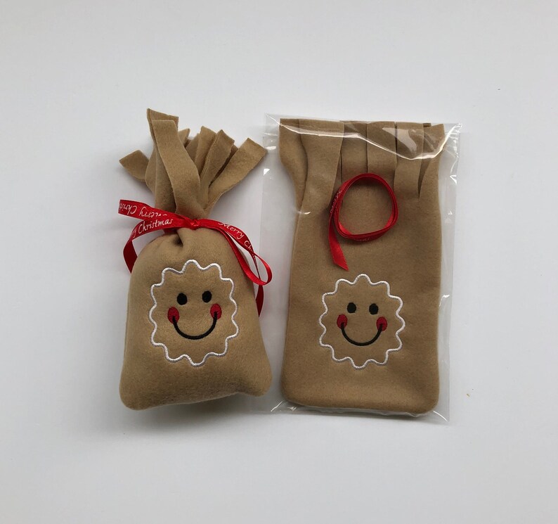 May include: Two brown felt gingerbread man ornaments with smiling faces. One ornament is in a bag with a red ribbon tied around it. The other ornament is in a clear plastic bag.