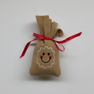 May include: A brown felt bag with a red ribbon tied around it. The bag has a white embroidered smiley face with red cheeks. The ribbon has the text "Happy Christmas" printed on it.