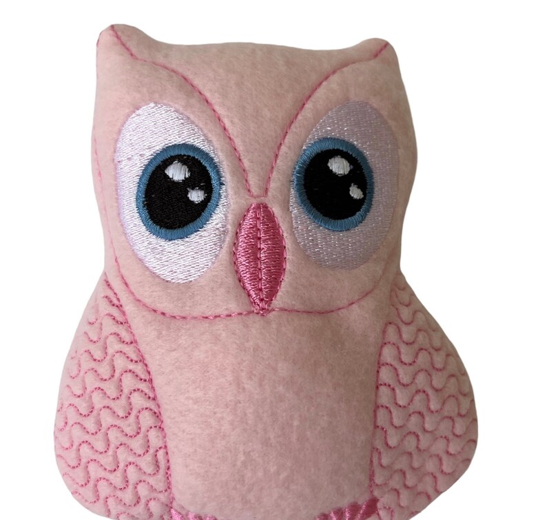 May include: A pink plush owl with embroidered eyes, beak, and feathers. The owl has a light blue and white eye with a black pupil.