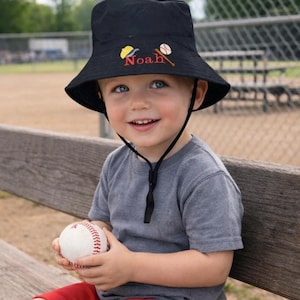 May include: A black bucket hat with the name "Noah" embroidered in red, along with baseball-themed graphics. The hat has a chin strap and is worn by a young child holding a baseball. The child is wearing a gray t-shirt and red shorts.