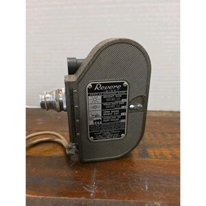 May include: Vintage Revere Eight movie camera in a gray textured finish. The camera has a lens, a handle, and a label with exposure guide information. The camera is sitting on a wooden surface.