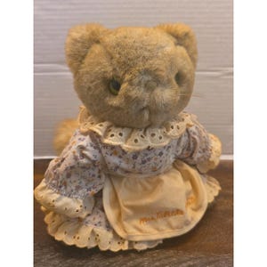 May include: A plush cat toy with tan fur and green eyes, wearing a white dress featuring a small floral pattern in purple and orange. The dress has a cream-colored eyelet lace collar and cuffs, and a cream apron with the text Mrs. Tabitha Twit.