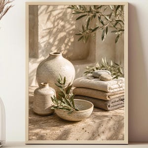 May include: A framed wall art print featuring a still life composition of two beige ceramic vases, a small bowl with olive branches, and a stack of folded neutral-toned towels, all set against a textured stone surface with soft shadows.
