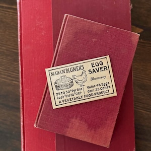 May include: A stack of two vintage red books with a small, aged paper label on top. The label reads "MADAM BLUMER'S EGG SAVER" with text about the product and a drawing of a basket of eggs and a chicken.