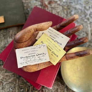 May include: A wooden hand sculpture holding several vintage church offering cards. The cards are yellow and white with handwritten text, including "Presbyterian Church" and the date "January 3, 1904." The hand rests on a red book.