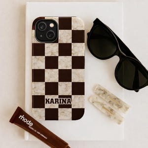 May include: A phone case with a brown and white checkered pattern and the name "KARINA" in bold, black letters. The image also includes black sunglasses, two marble hair clips, and a tube of brown lip product.