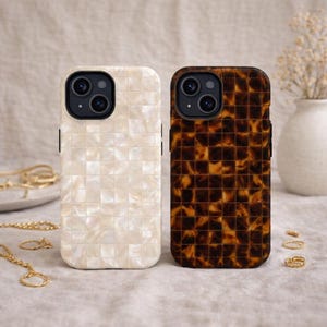 May include: Two phone cases with a square pattern. One case is off-white, the other is brown with a tortoiseshell design. Both cases have a black camera module. Gold jewelry and a vase of flowers are in the background.