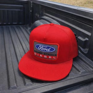May include: A red baseball cap with a flat brim. The cap features a rectangular patch with the Ford logo in blue and white, and the words "POWER STROKE DIESEL" in red.