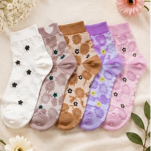 May include: Five pairs of ankle socks in white, mauve, tan, lavender, and light pink. Each sock features a textured, raised floral pattern across the fabric and small, contrasting embroidered flower accents in black, green, or yellow.