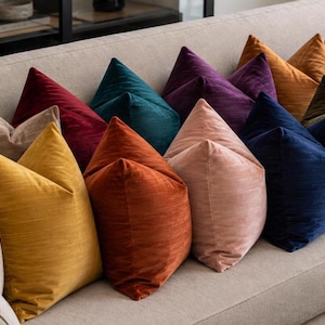 Peut inclure: Une collection de coussins carrés dans une variété de couleurs unies, notamment crème, jaune, orange, rose, rouge, bleu sarcelle, violet, bleu, vert olive et gris anthracite, avec une finition en tissu doux et texturé sur un canapé beige.