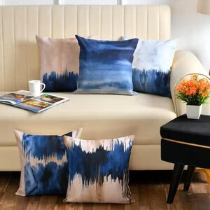 May include: A set of five square throw pillows featuring a blue and beige abstract watercolor pattern. Three pillows are arranged on a cream-colored sofa, while two are positioned on the floor. The designs display varying gradients of blue.