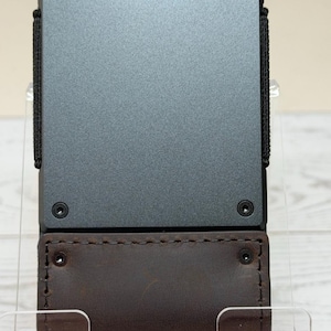 May include: A modern wallet with a dark gray metal body and brown leather accents. The wallet features a black elastic band and is held together with small screws. The leather section is approximately 3 inches in height.
