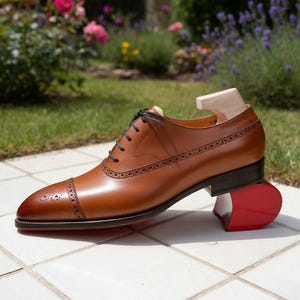 May include: A brown leather Oxford shoe with decorative perforations and dark brown laces. The shoe is on a red shoe tree and is set against a background of flowers and greenery.