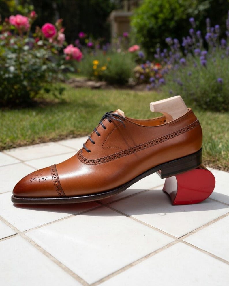 May include: A brown leather Oxford shoe with decorative perforations and dark brown laces. The shoe is on a red shoe tree and is set against a background of flowers and greenery.