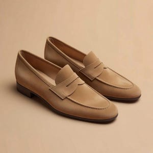 May include: A pair of tan suede penny loafers featuring a classic moc-toe construction, a horizontal strap across the vamp with a central cutout, a low stacked heel, and a smooth interior lining, presented in a clean, uniform light brown color.