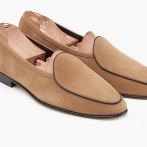May include: A pair of tan suede loafers with black trim and soles. The shoes have a classic slip-on design and are shown with wooden shoe trees inside. The loafers are a neutral color and suitable for various outfits.