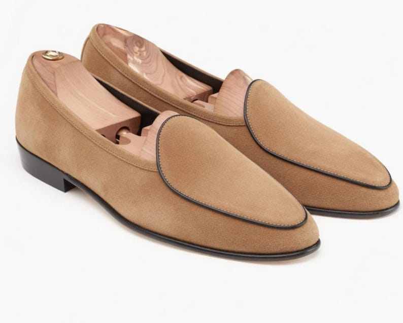 May include: A pair of tan suede loafers with black trim and soles. The shoes have a classic slip-on design and are shown with wooden shoe trees inside. The loafers are a neutral color and suitable for various outfits.