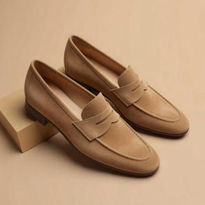May include: A pair of tan suede loafers featuring a classic penny slot design across the vamp, a rounded toe, and a low heel. The shoes are crafted with a smooth, uniform texture and a clean silhouette, showcasing a slip-on style with tonal stitching.