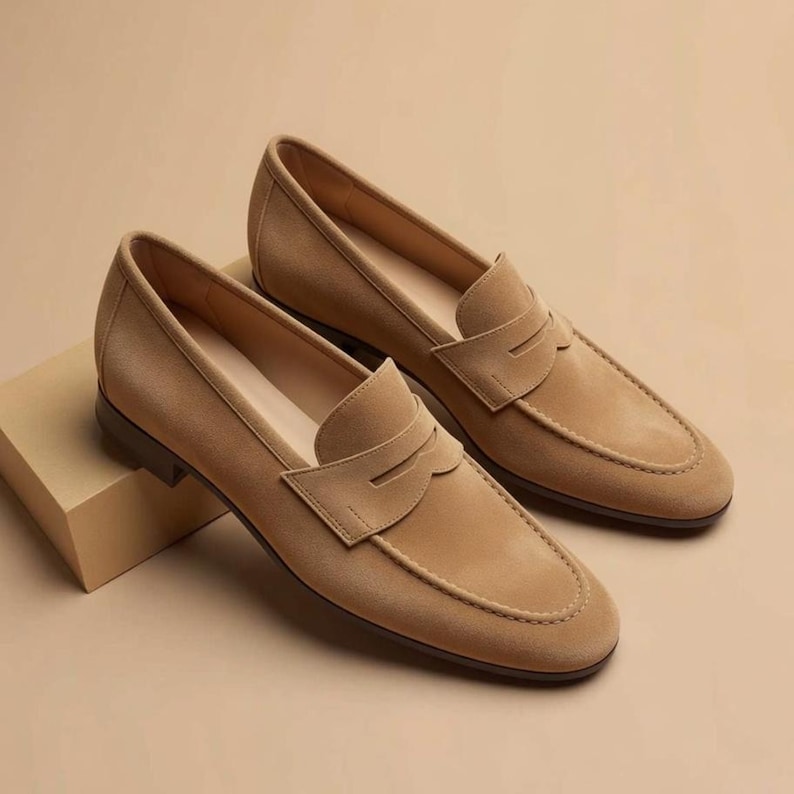 May include: A pair of tan suede loafers featuring a classic penny slot design across the vamp, a rounded toe, and a low heel. The shoes are crafted with a smooth, uniform texture and a clean silhouette, showcasing a slip-on style with tonal stitching.