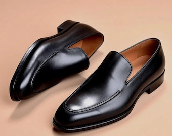 May include: A pair of black leather loafers featuring a smooth, polished finish, a rounded toe, a low heel, and a tan interior lining. The slip-on shoes have a clean, minimalist design with visible stitching along the top vamp and a structured silhouette.
