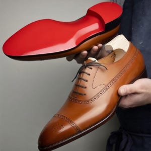 May include: A pair of brown leather dress shoes with a red sole. The shoes have a classic design with intricate stitching and a cap toe. The sole is a vibrant red color, and the shoes are held up in the air.