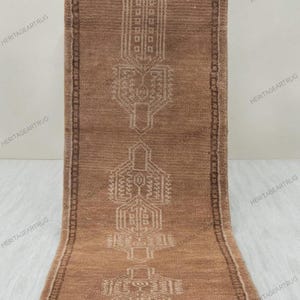 May include: A rectangular runner rug in a medium brown tone featuring a series of three symmetrical, geometric white line patterns centered along the length. The rug has a dark brown border with a repeating pattern and white fringe at both ends.