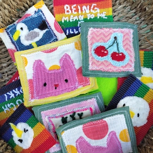 May include: A collection of small, square fabric patches featuring embroidered designs including a seagull, a pink cat, red cherries, and a rainbow background with the text "BEING MEAN TO ME IS ILLEGAL". The patches have stitched edges.
