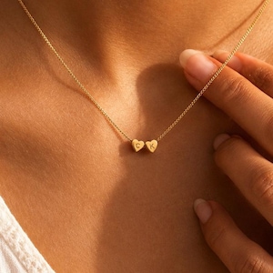 May include: A gold-toned necklace featuring a delicate chain and two small heart-shaped charms. Each heart charm is engraved with a single letter, specifically the letters C and K, positioned side by side at the center of the chain.