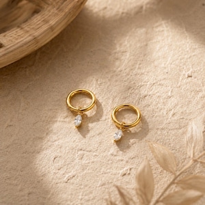 May include: A pair of gold hoop earrings, each featuring a single dangling marquise-cut clear gemstone. The earrings have a polished metallic finish and a simple circular hoop design that supports the small, faceted stone charm hanging from the base.