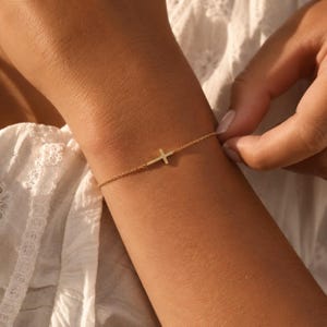 May include: A delicate gold chain bracelet featuring a small, centered horizontal cross charm. The bracelet is constructed from fine, polished gold links that wrap around the wrist, showcasing a minimalist design with a simple, clean metal finish.