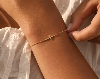 May include: A delicate gold chain bracelet featuring a small, centred horizontal cross charm. The bracelet is constructed from fine, polished gold links that wrap around the wrist, showcasing a minimalist design with a simple, clean metal finish.