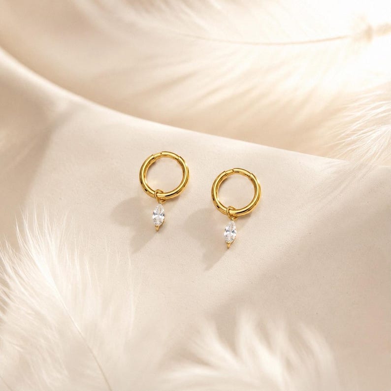 May include: A pair of gold hoop earrings, each featuring a single marquise-cut clear gemstone charm suspended from the bottom of the hoop. The earrings are crafted from a polished gold-tone metal, providing a bright and reflective finish to the design.