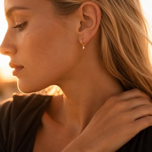 May include: A small gold hoop earring featuring a single, delicate marquise-cut clear gemstone charm suspended from the bottom. The earring is worn in the earlobe, showcasing a minimalist design with a polished metallic finish and a clear stone.