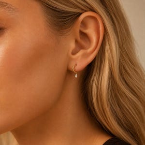May include: A small gold hoop earring featuring a single, delicate marquise-cut clear gemstone charm suspended from the bottom. The earring is worn in the earlobe, showcasing a polished metallic finish and a minimalist, refined design aesthetic.
