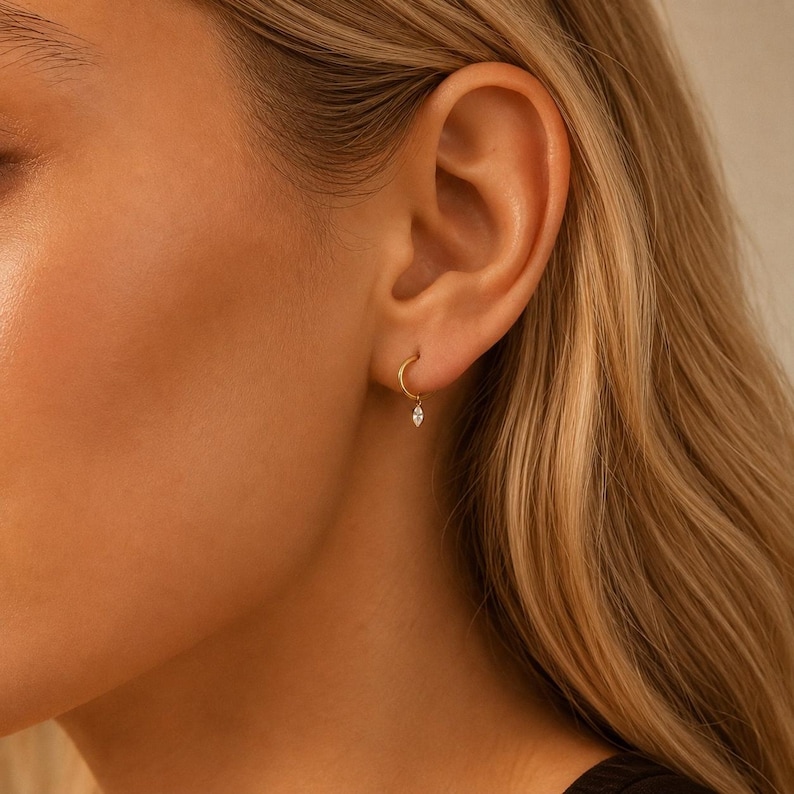May include: A small gold hoop earring featuring a single, delicate marquise-cut clear gemstone charm suspended from the bottom. The earring is worn in the earlobe, showcasing a polished metallic finish and a minimalist, refined design aesthetic.