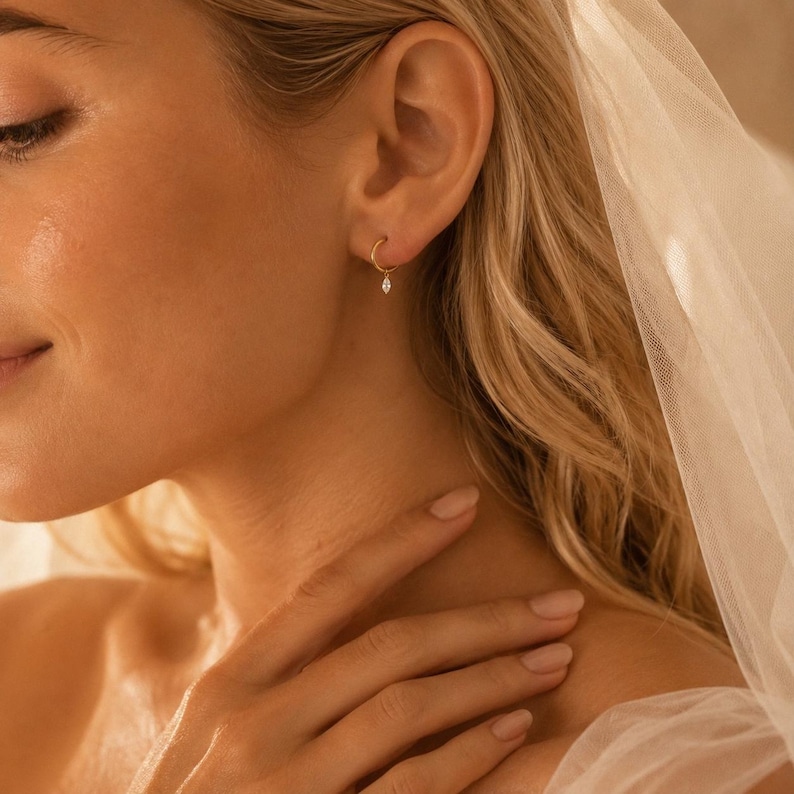 May include: A small gold hoop earring featuring a single marquise-cut clear gemstone charm suspended from the bottom. The earring is worn in the earlobe, showcasing a delicate metallic band and a faceted stone that catches the light.