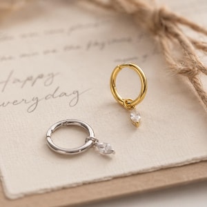 May include: Two small hoop earrings, one in gold tone and one in silver tone, each featuring a single marquise-cut clear gemstone charm dangling from the bottom. The hoops have a hinged closure mechanism and a smooth, polished metallic finish.