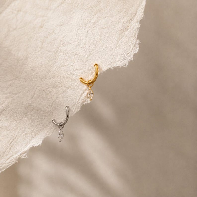 May include: Two small hoop earrings, one in gold tone and one in silver tone, each featuring a single marquise-cut clear gemstone charm dangling from the bottom of the hoop. The earrings are positioned against a textured, off-white paper edge.