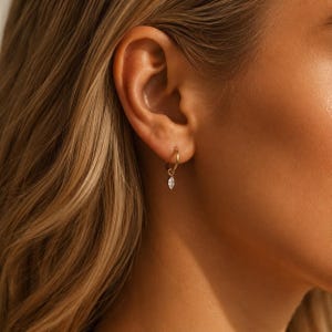 May include: A gold hoop earring featuring a small, dangling marquise-cut clear gemstone. The hoop is thin and circular, securing the faceted stone which hangs vertically from the bottom of the metal ring, creating a delicate and simple jewellery piece.