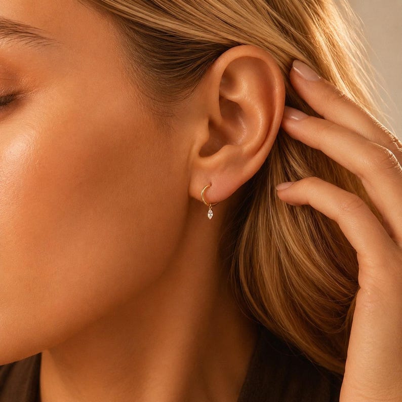 May include: A small gold hoop earring featuring a single marquise-cut clear gemstone charm dangling from the bottom. The hoop is thin and circular, crafted from polished yellow gold, and the gemstone is set in a delicate metal frame.