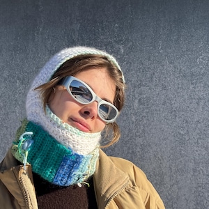 May include: A knitted balaclava in white, teal, and blue, with a matching scarf. The balaclava is worn with silver-framed sunglasses and a tan jacket. The knitted hood covers the head and neck, offering warmth and style.