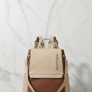 May include: A two-tone backpack in beige and brown leather. The bag has a flap closure with a zipper pocket, adjustable shoulder straps, and a detachable shoulder strap. The background is a white marble wall.