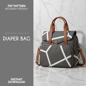 Op de afbeelding: Grijze luiertas met een wit geometrisch patroon en bruine leren handvatten en accenten. De tas heeft een schouderriem en een voorvak. Tekst op de tas zegt "DIAPER BAG" en "PDF PATTERN BEGINNER FRIENDLY".