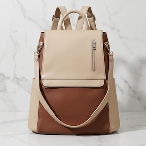 May include: A two-tone backpack in beige and brown leather. The top flap and straps are beige, while the main body is brown. Features include a top handle, adjustable straps, and a front zipper pocket. The bag has a sleek, modern design.