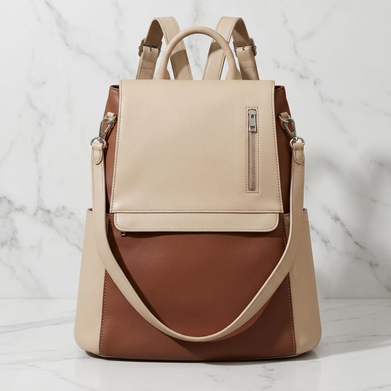May include: A two-tone backpack in beige and brown leather. The top flap and straps are beige, while the main body is brown. Features include a top handle, adjustable straps, and a front zipper pocket. The bag has a sleek, modern design.