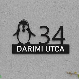 Pode incluir: Placa de endereço de metal preto com um design de pinguim, o número 34 e o texto "DARIMI UTCA" em branco. A placa é montada em uma parede cinza texturizada, com vegetação ao fundo.